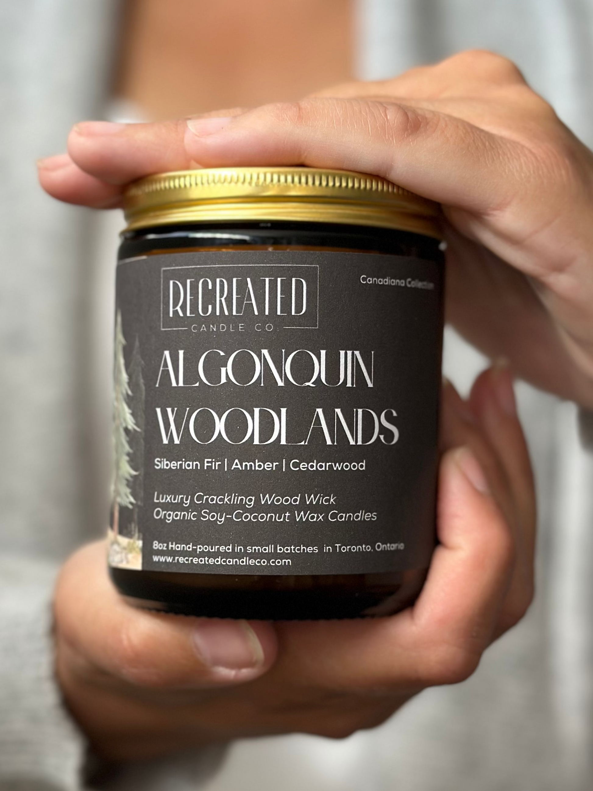 Person holding a candle jar labeled 'Regreated Algonquin Woodlands' against a neutral background